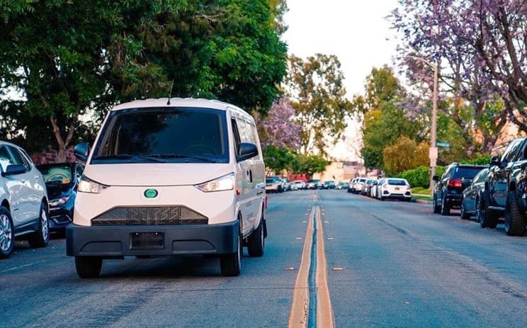 White EV van driving toward camera along shaded street.
