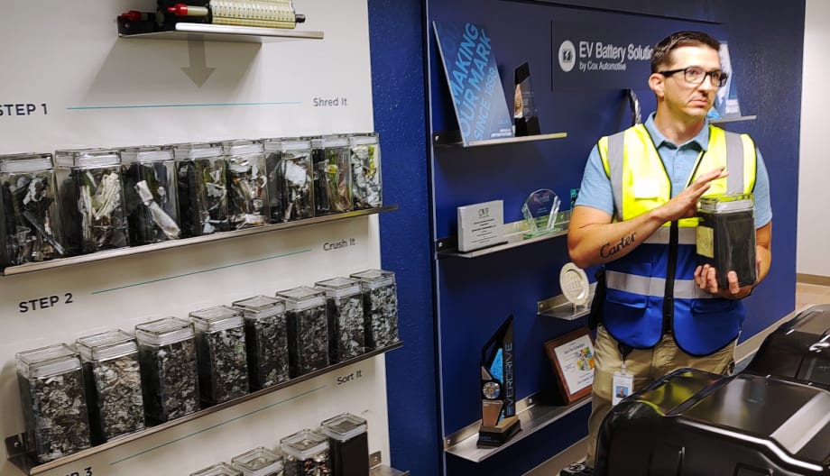 A man dressed in a safety vest is holding clear container of fine particles of black mass.
