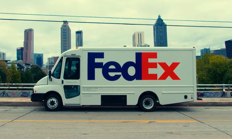 White battery-electric FedEx truck parked in front of a city skyline.