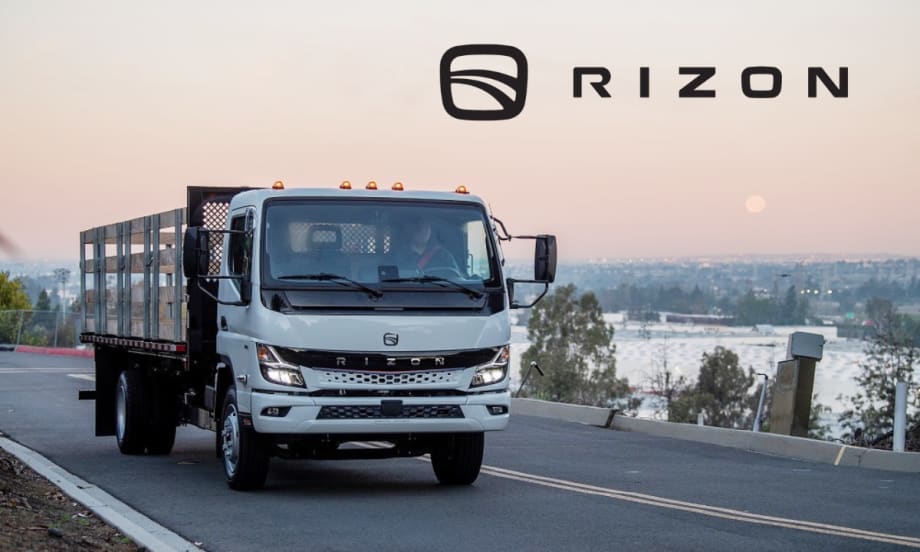 Stake body medium-duty electric truck driving on a road with sunset in background and Rizon logo upper right against sky.