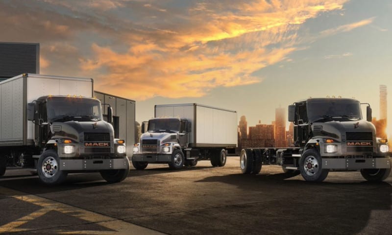 Three Mack electric trucks parked in front of a scenic afternoon sky.