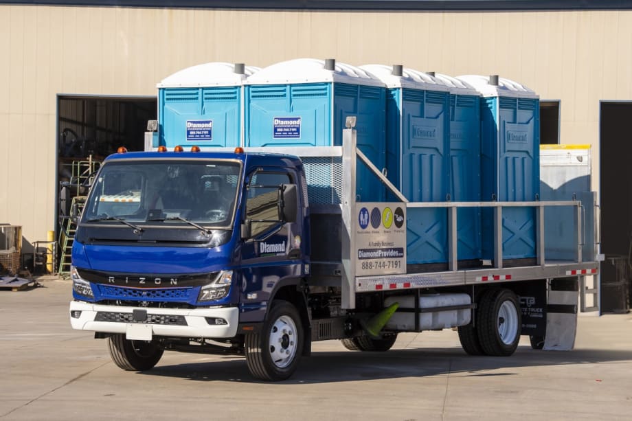 Battery electric truck with blue cab and flatbed hauling portable toilets.