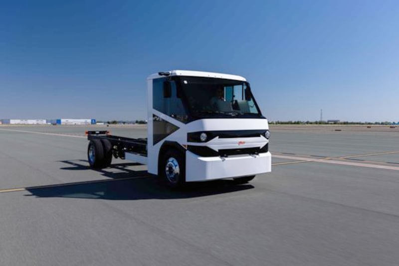 White battery-electric chassis cab parked in open paved area with blue sky.