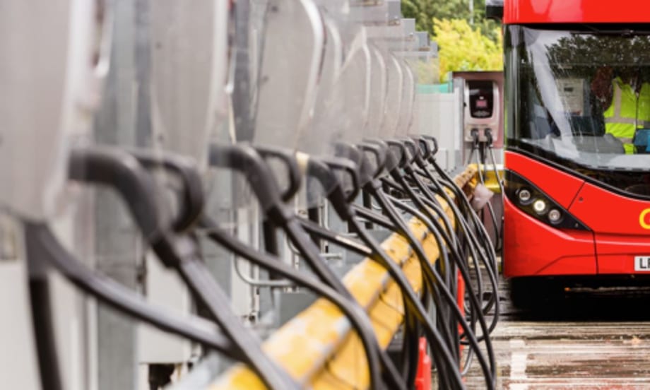 A row of electric bus charging stations with charging cables lined up, with a red double-decker bus plugged in and charging at the end of the row.