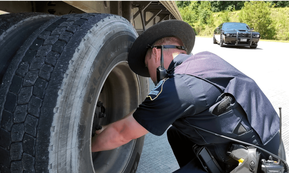 A state police officer inspects the wheels of a trailer with his patrol car parked in the background.