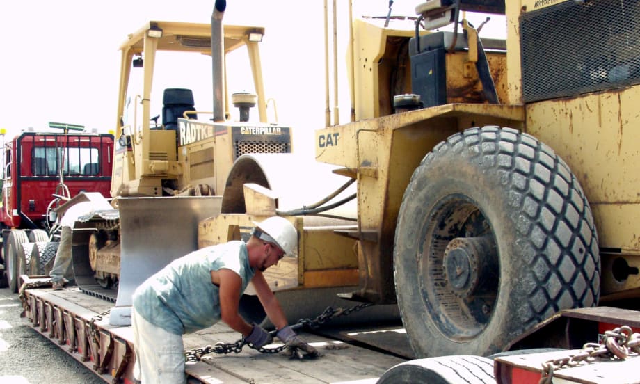 Construction worker tightens chain to secure a large piece of equipment on a trailer.