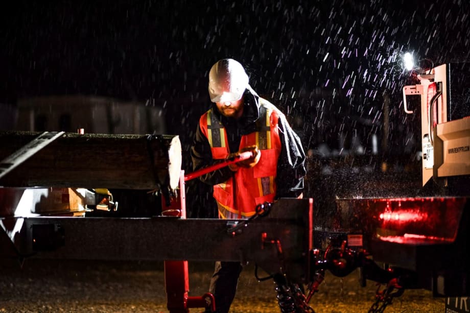 Worker wearing a high-visibility jacket and hard hat operates heavy equipment during a nighttime storm, with rain pouring down and debris secured on a trailer.