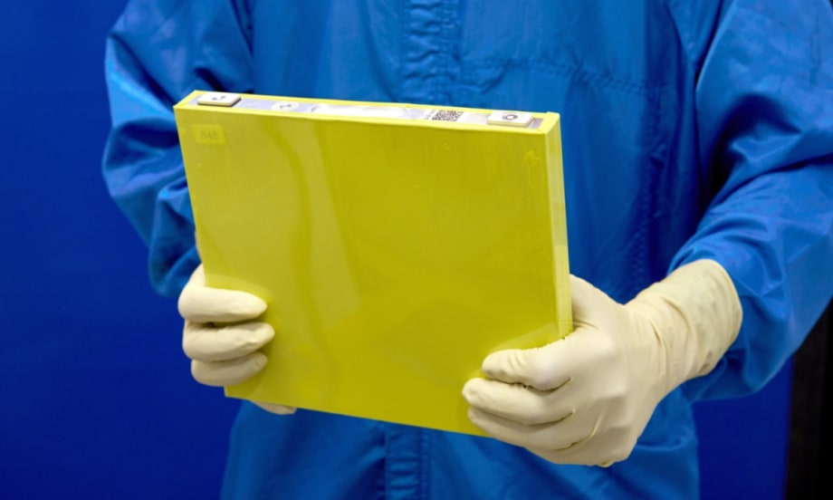 Closeup of person in blue lab clothing wearing gloves and holding a yellow battery cell.