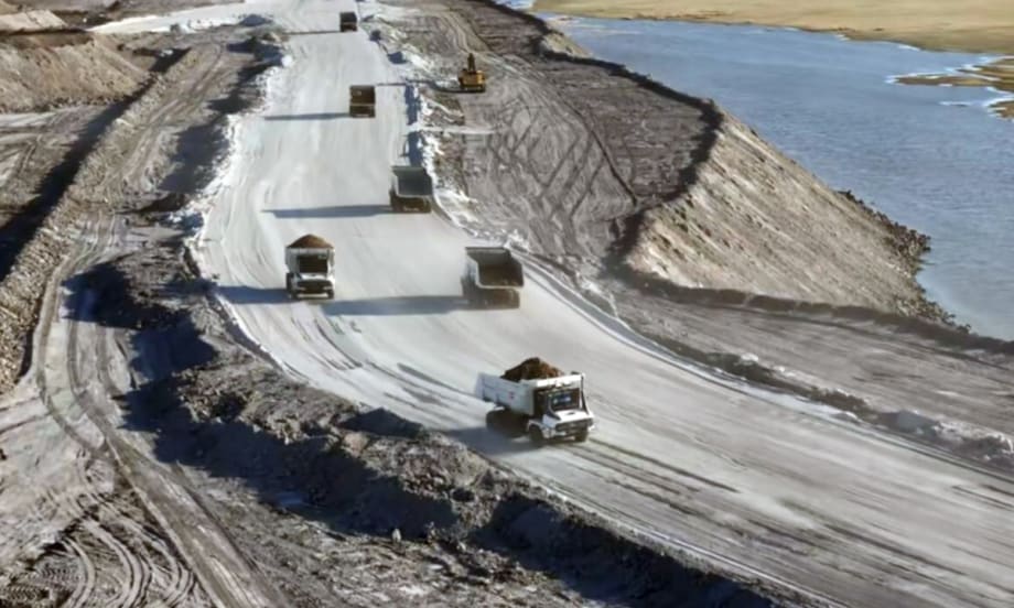 View of mining dump trucks driving on a curved road on top of a hill next to water.