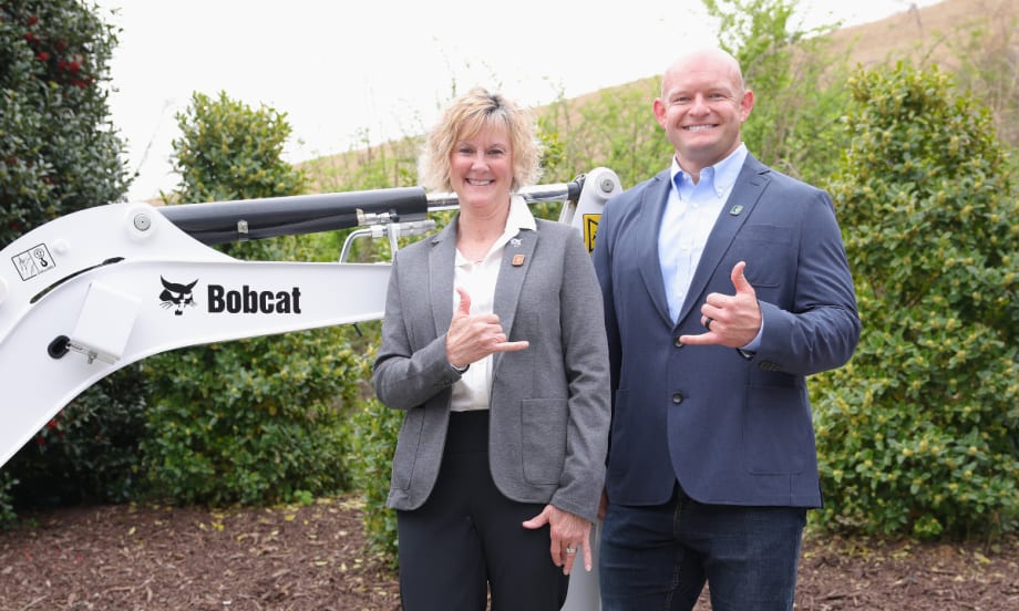 A woman and a man in business attire pose in front of a Bobcat machine, smiling and making shaka hand gestures.