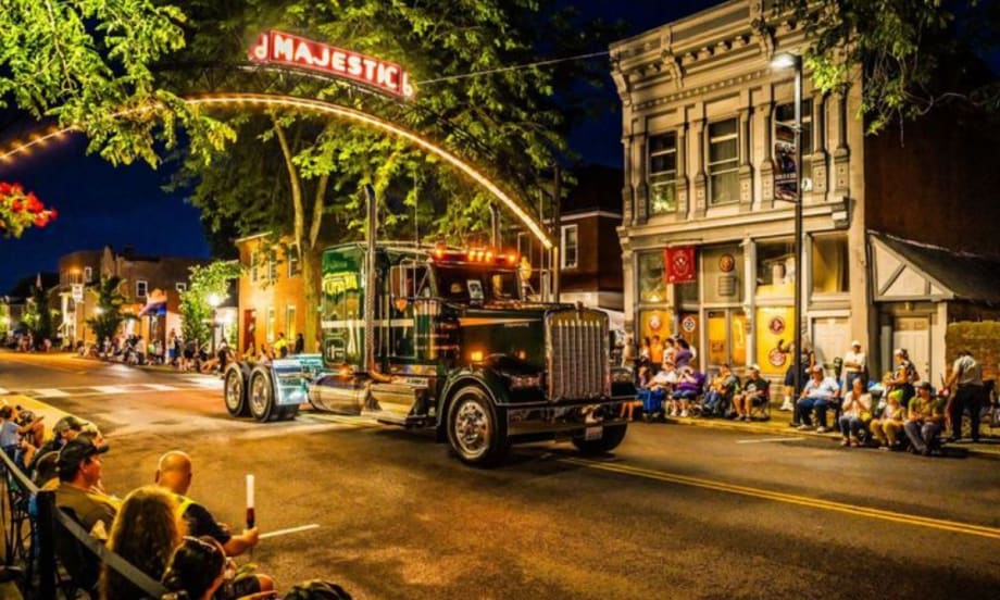 A classic Kenworth truck drives under a lit "Majestic" sign during the nighttime Kenworth Truck Parade in downtown Chillicothe, Ohio, as crowds line the street to watch the event.