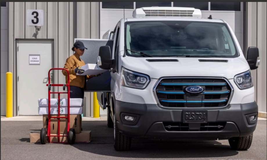 A delivery worker unloads boxes from a Ford electric refrigerated van parked outside a cold storage facility, illustrating electric truck use in commercial fleet operations.