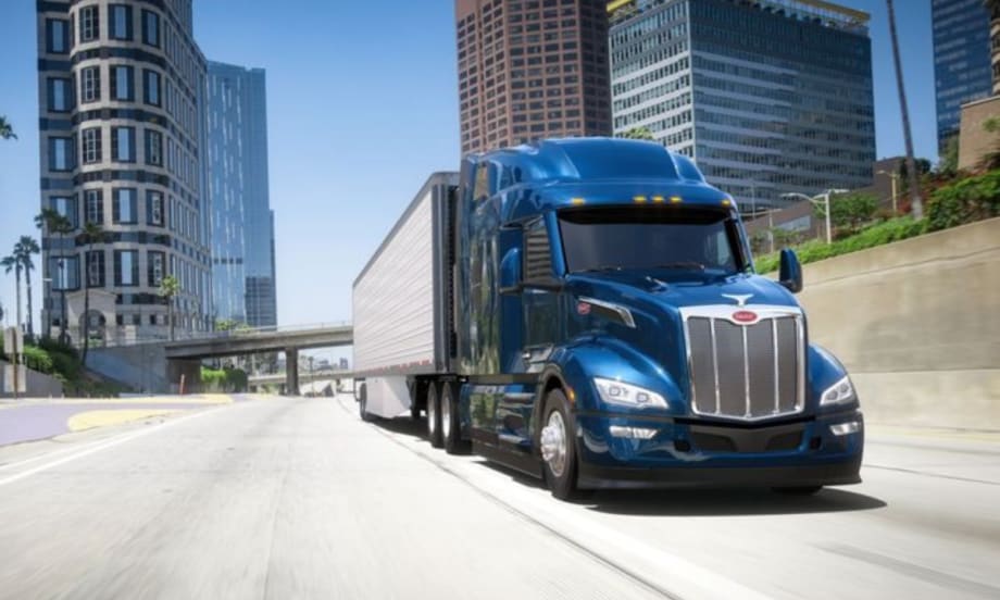 A blue Peterbilt Model 579 semi-truck driving on a city highway with tall buildings in the background.