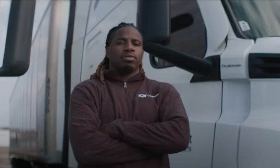 A truck driver stands with arms crossed in front of a white semi-truck, wearing a maroon jacket, featured in the ATA’s DRIVEN series highlighting professional drivers in the trucking industry.