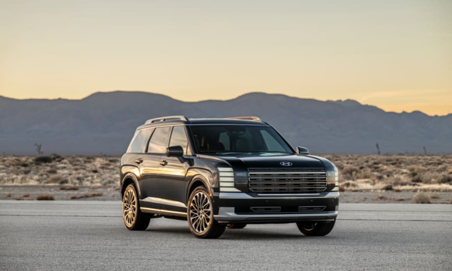 Front three-quarter view of the 2026 Hyundai Palisade SUV parked on a desert road at sunset, with mountains in the background.