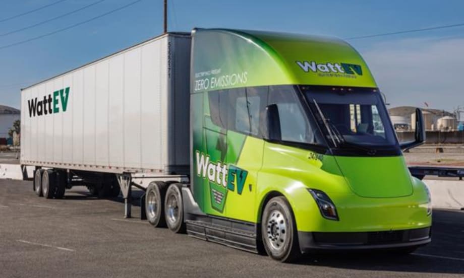 A WattEV electric semi-truck with a green cab and white trailer is parked in an industrial area under clear skies, showcasing the company's zero-emissions freight branding.