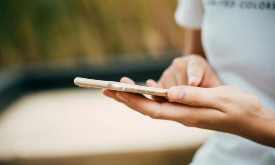 Close-up of a person using a smartphone, holding it with one hand and tapping the screen with the other, wearing a white shirt with blurred outdoor background.