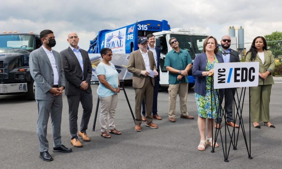 Group of officials standing around woman making announcement with two battery-electric trucks in the background.