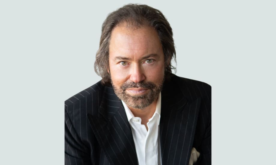 A man in a pinstripe suit jacket and white collared shirt looks directly at the camera with a neutral expression, seated against a light gray background.