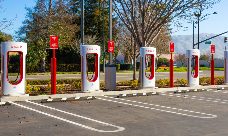 Row of Tesla Supercharger stations in a parking lot, ready for electric vehicles to charge, with clear skies and mountains visible in the background.