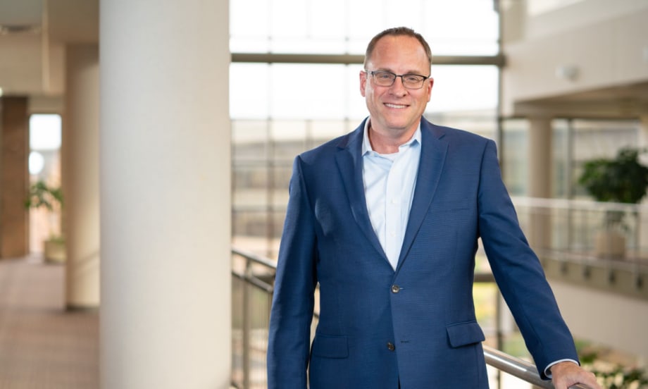 A man in a blue suit and glasses stands indoors near a railing, smiling, with large windows and natural light in the background.