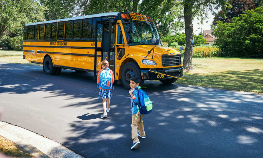 Two children with backpacks walk toward a First Student yellow school bus parked on a suburban street under the shade of trees.
