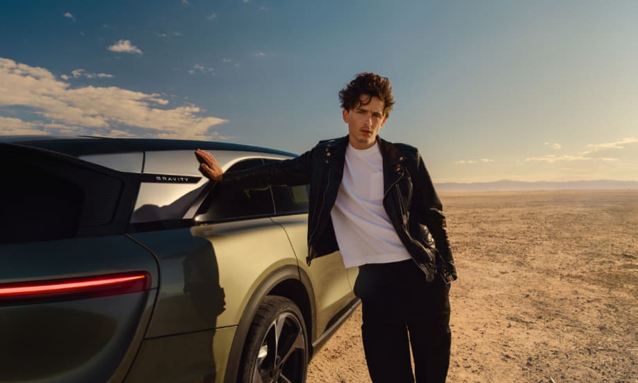 Actor Timothée Chalamet leans against a Lucid Gravity SUV in a desert setting, wearing a black leather jacket and white shirt, as part of Lucid’s new marketing campaign.