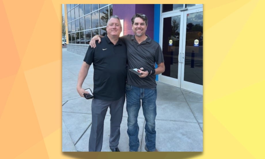 Two men stand smiling outside a building with glass doors, one with his arm around the other. Both are holding phones and dressed in casual collared shirts, one in black and one in gray.