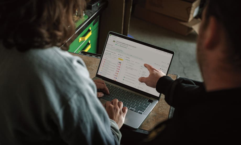 Two people review John Deere’s Operations Center PRO Service on a laptop, showing diagnostic trouble codes and repair tools for equipment maintenance.