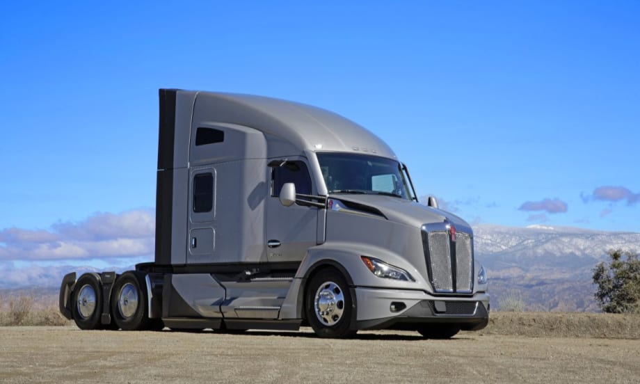 A Kenworth T680 tractor-trailer with a 76-inch sleeper cab parked on a dirt lot with mountains in the background under a clear blue sky.