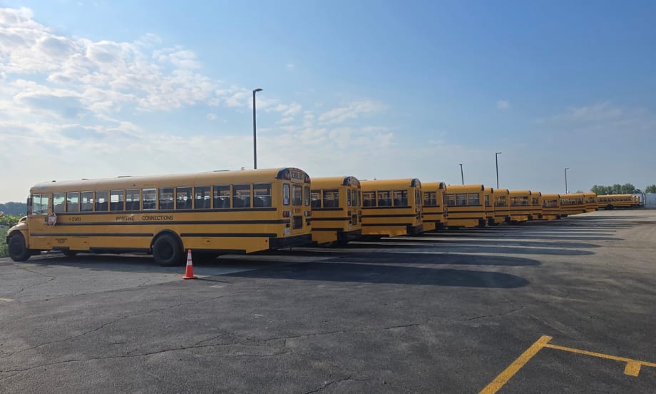 Row of yellow International CE Series electric school buses parked in a lot in Bloom Township, Illinois, ready for deployment. One bus is marked “EV01” with an orange cone nearby.