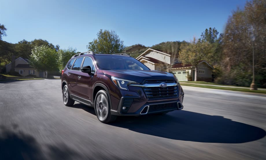 A maroon 2026 Subaru Ascent three-row SUV with a bold black front grille, LED headlights, and roof rails drives through a suburban neighborhood on a sunny day.