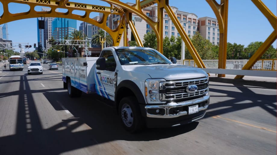 A white service truck driving across a yellow bridge with city skyline in the background.