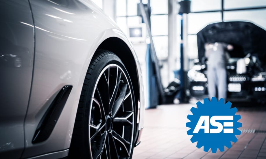 Close-up of a car wheel in an auto repair shop with a mechanic working in the background; ASE logo displayed in the foreground.