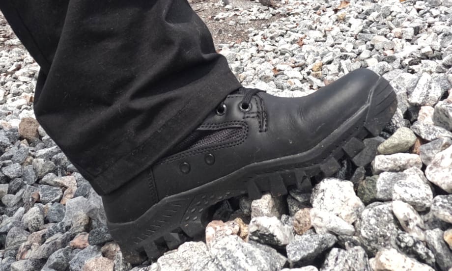 Black tactical boot with rugged sole standing on gray stones and gravel