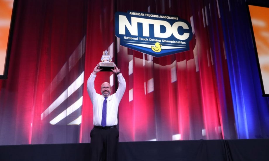 David Comings holds the NTDC Grand Champion trophy above his head on stage at the 2025 ATA National Truck Driving Championships.