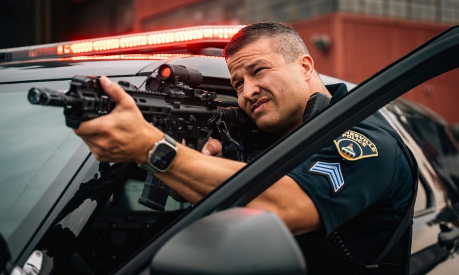 Police officer aiming tactical rifle from patrol vehicle with emergency lights