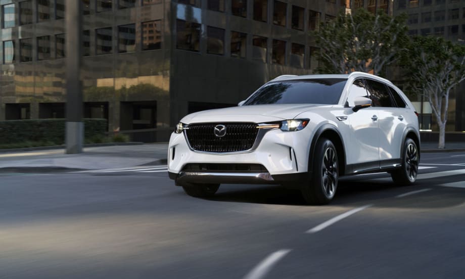 A white 2026 Mazda CX-90 crossover SUV driving through a city street, showcasing its front grille, LED headlights, and modern exterior design.