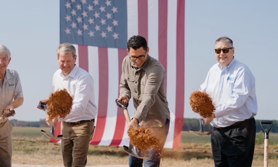 Rivian CEO RJ Scaringe, Georgia Governor Brian Kemp, and other officials shovel dirt during the groundbreaking ceremony, with a large American flag in the background.