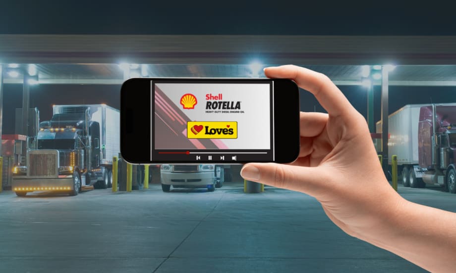 Hand holding a smartphone displaying a Shell Rotella and Love’s Travel Stops video screen, with semi-trucks parked at a brightly lit fuel station in the background.