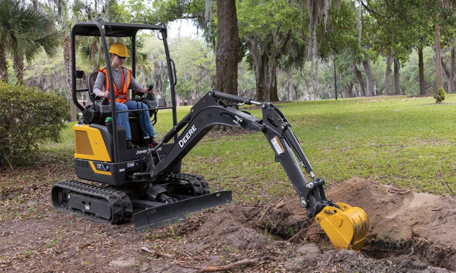 Operator using a John Deere 17 P-Tier compact excavator to dig in a grassy park area surrounded by trees.