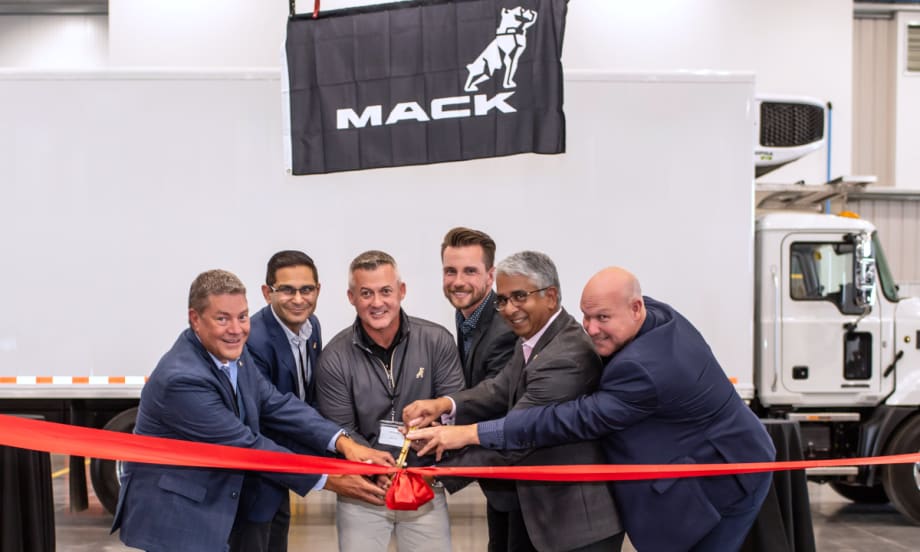 Group of businessmen cut ribbon in front of a white box truck with a Mack Trucks sign on the side.