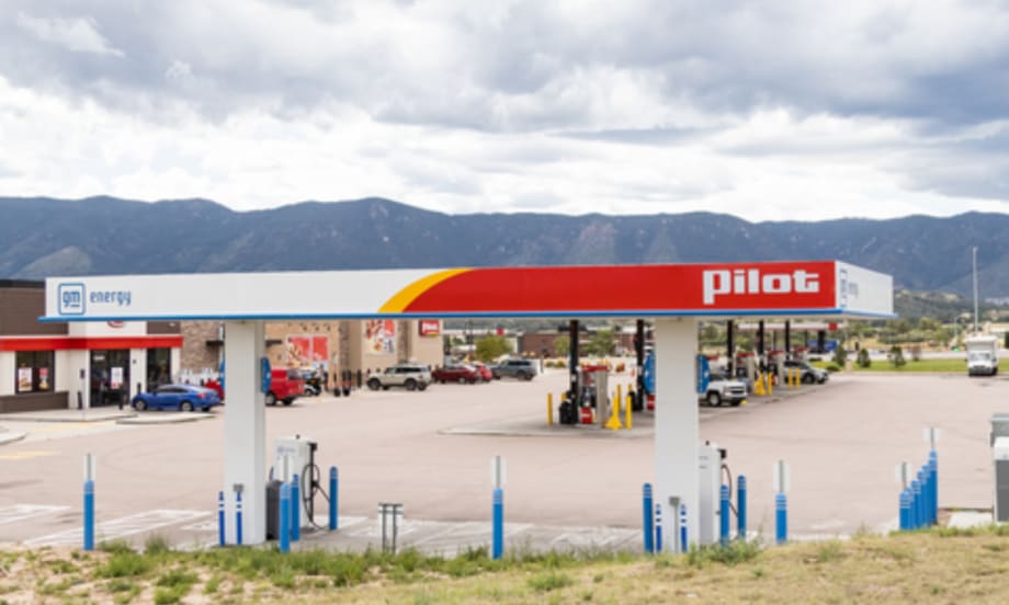 A Pilot Travel Center with GM Energy and EVgo fast-charging stations installed under the canopy, with fuel pumps and vehicles in the background against a mountain landscape.