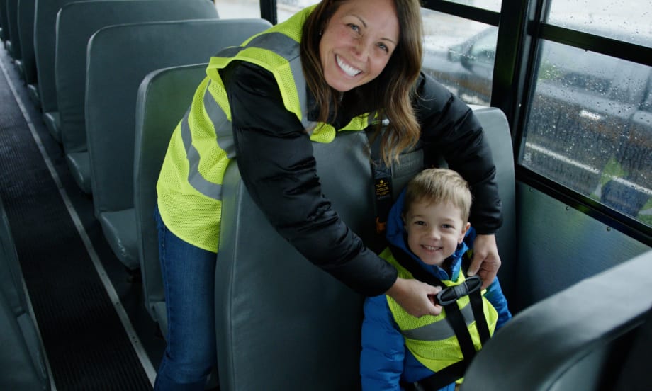 A school bus driver helps a young student buckle up while smiling inside a bus, reflecting First Student’s First Serves behavioral support program.