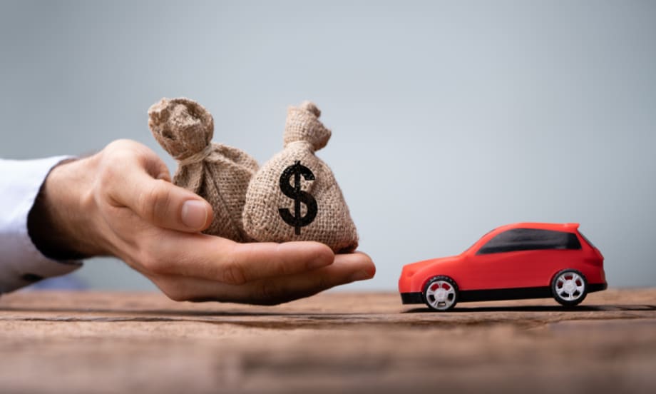 A hand holding small burlap money bags next to a toy red car, symbolizing auto financing, loan payments, and dealership profitability.