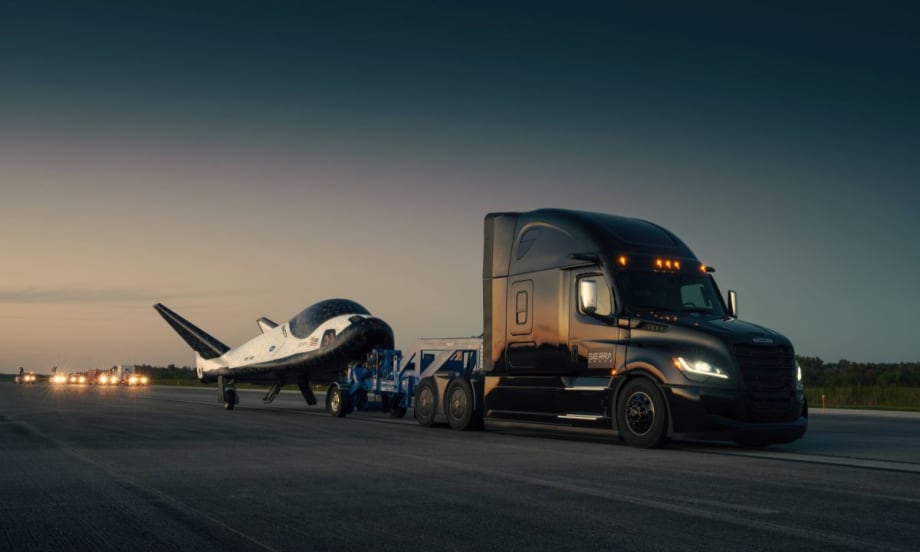 A Freightliner Cascadia semi-truck towing Sierra Space’s Dream Chaser spaceplane on a runway at dusk.