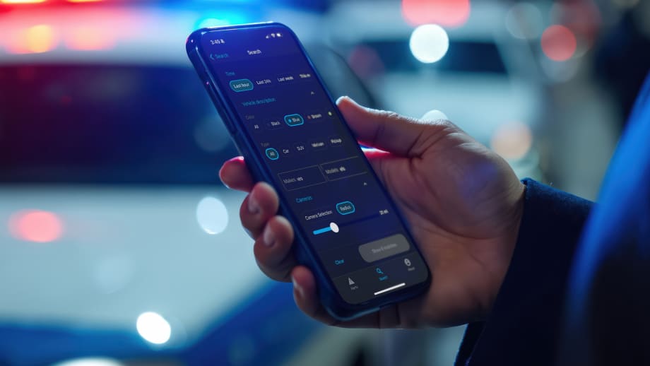 Closeup photo of man's hand holding a phone with an app displayed and out of focus police cars in the background