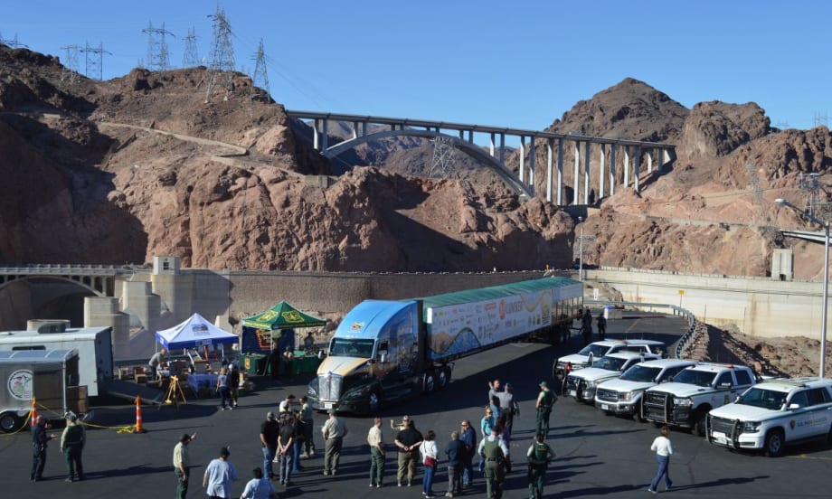 A semi-truck hauling the U.S. Capitol Christmas Tree trailer is parked near Hoover Dam, with people and law enforcement vehicles gathered nearby.