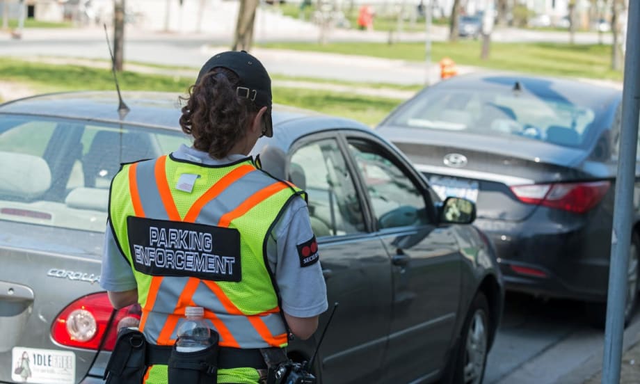 woman officer shown from the back in a vest that states parking enforcement while issuing a parking ticket to a car