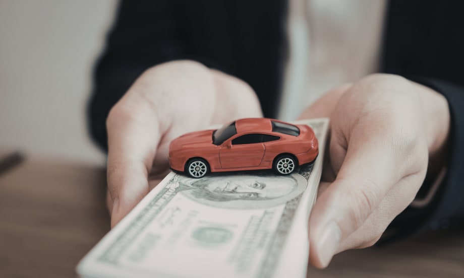 A person holds a stack of cash with a small red toy car on top.